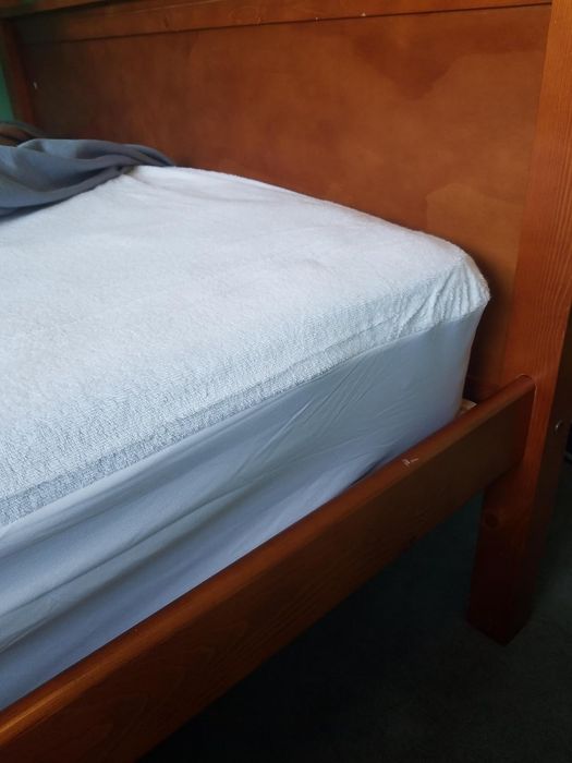 A close-up shot of a well-made bed featuring a white mattress protector, complemented by a wooden bed frame. A grey blanket partially drapes over the mattress