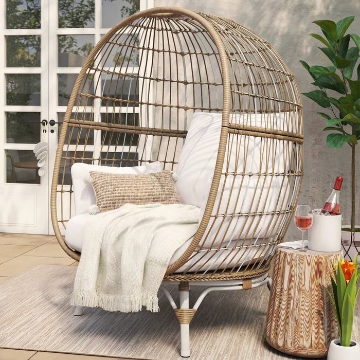Egg-shaped rattan chair with soft white cushions and a cozy blanket, placed outdoors beside a small table with a wine glass and bottle