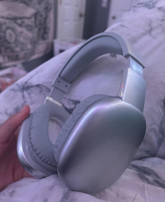 A close-up shot of someone's hand holding silver over-ear headphones, resting on a bed with a patterned blanket in the background.