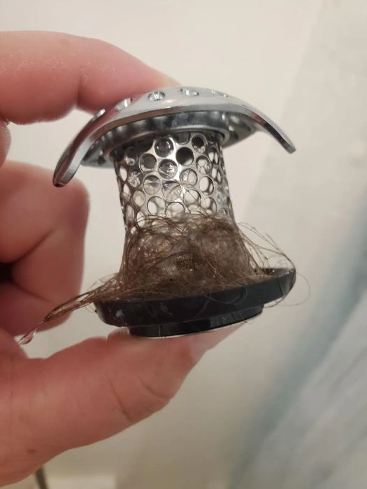 A hand holding a metal drain hair catcher, with hair tangled around it