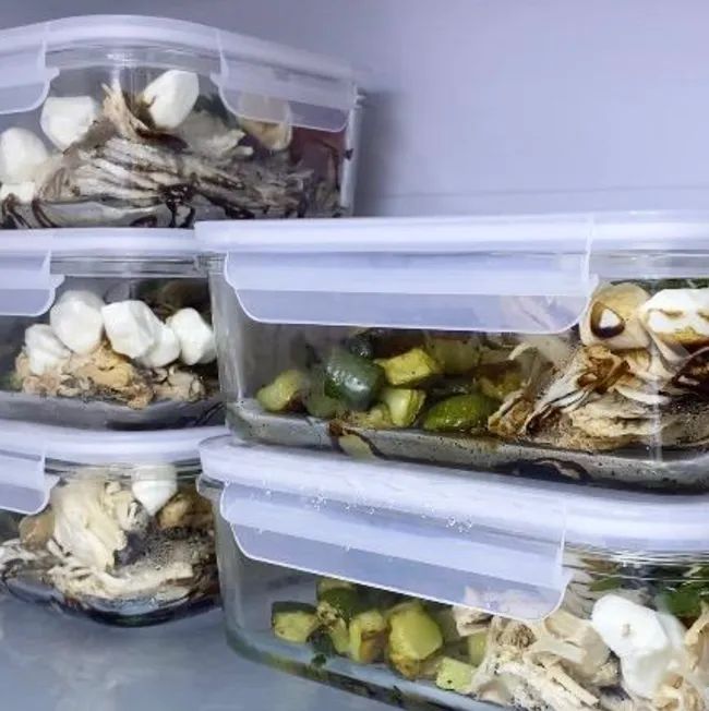 Five clear square meal prep containers with white snap-on lids, neatly storing prepped meals in a fridge.