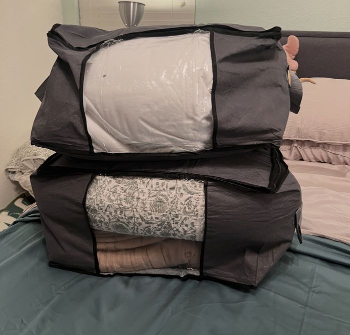 Two large, grey fabric storage bags packed with neatly folded bedding, placed on a bed adorned with pink and grey sheets