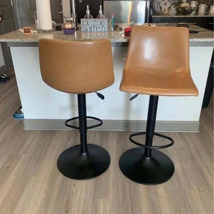 Two contemporary bar stools with tan upholstered seats and sleek black bases, perfectly complementing a modern kitchen aesthetic. A countertop with decorative elements is visible in the background.