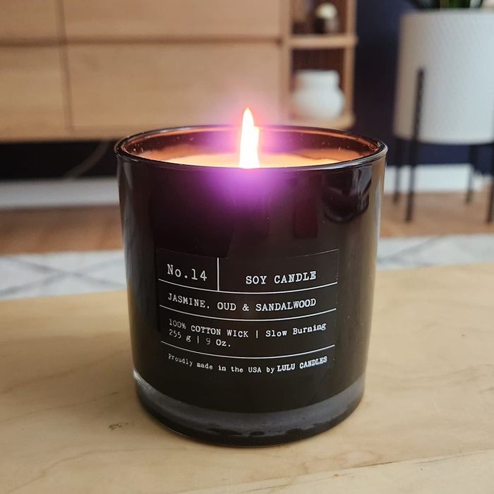 A lit candle in a black glass container with white text, indicating a jasmine, oud, and sandalwood fragrance and a cotton wick.