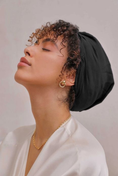 A person with curly hair wearing a silk turban and hoop earrings, their eyes gently closed and head tilted up in serenity
