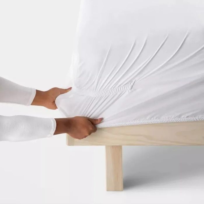 A person securing a white fitted sheet on a bed, making sure the corner seam fits snugly