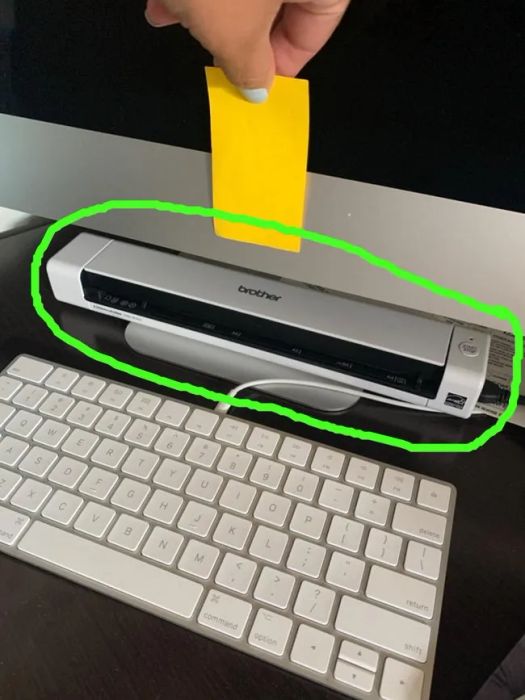 A hand holds a yellow sticky note above a Brother scanner, which is placed on top of a keyboard