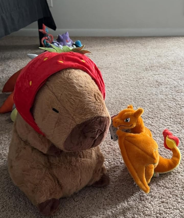 A plush capybara wearing a red bandana and a tiny yellow dragon toy, sitting on a soft carpet.