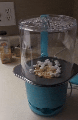 Popcorn popping inside a blue air popper in a kitchen, with kernels visible and kitchen counter items in the backdrop, including electrical outlets.