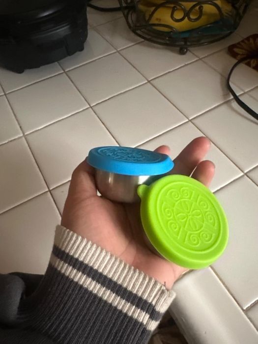 A person holding two small stainless steel bowls, each with blue and green silicone lids, on a kitchen counter
