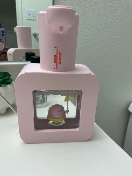 A pink soap dispenser with a clear window revealing a dinosaur figure inside