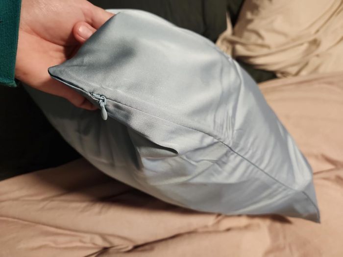 A close-up of a hand holding a satin pillowcase with a zipper resting on a bed.