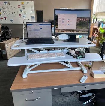 A white, two-tier desk riser with a keyboard on the lower shelf and a laptop and monitor positioned on the higher shelf.