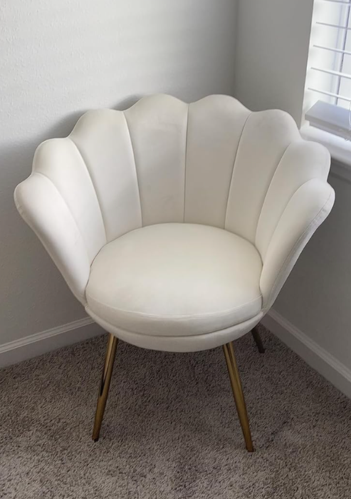 A white upholstered scalloped chair with gold legs, elegantly positioned in the corner of a room with plush carpeting