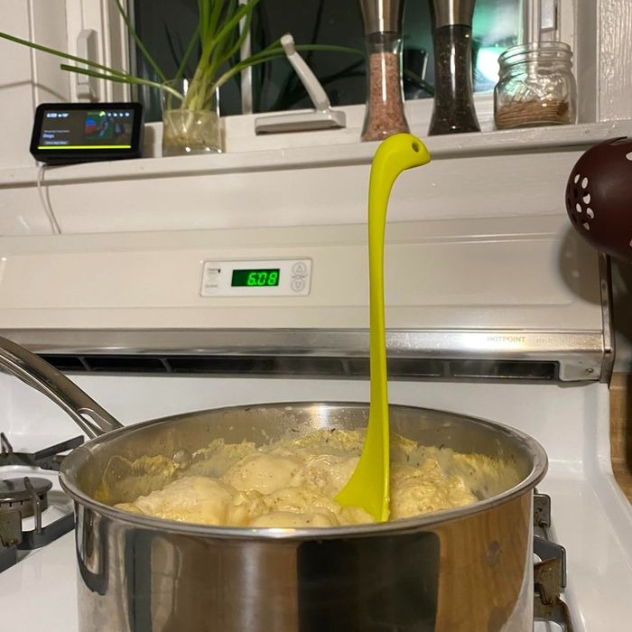 A cozy kitchen setup with a pot simmering on the stove; inside, a dinosaur-shaped ladle stands tall. Nearby, spices and a smart speaker complete the scene.