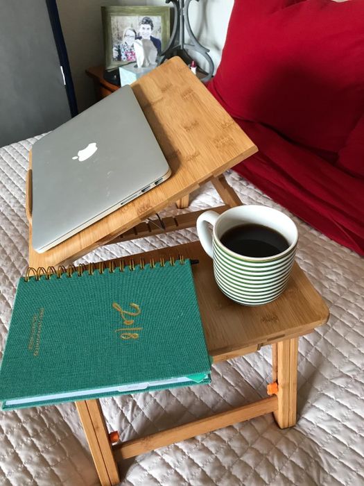 A laptop desk on a bed with a laptop, green notebook, and a striped cup of coffee. The cozy bedroom setting creates the perfect work environment