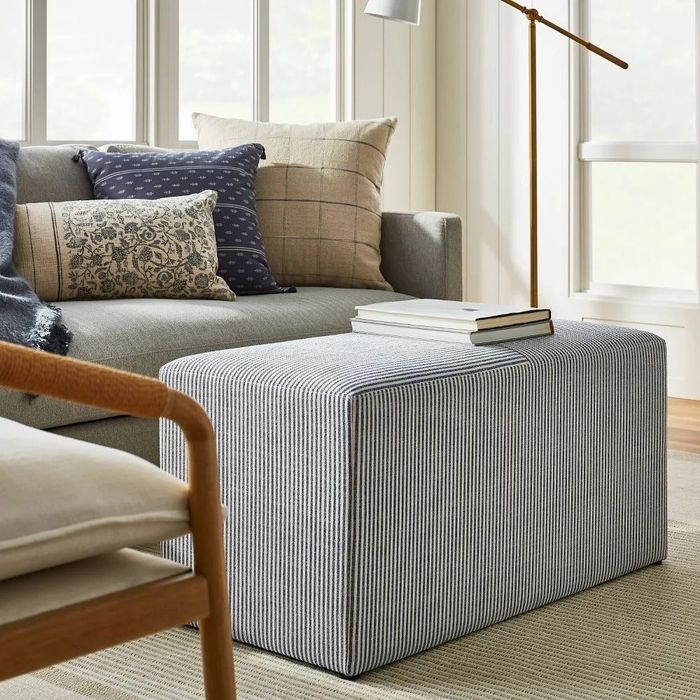 Minimalist living room featuring a gray sofa, patterned throw cushions, striped ottoman, and floor lamp. Books are stacked on the ottoman in a cozy and chic arrangement