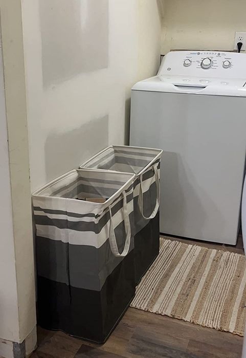 A well-organized laundry room featuring a washing machine, a dual-compartment striped laundry hamper, a cozy rug, and shelves adorned with decorative and household essentials.