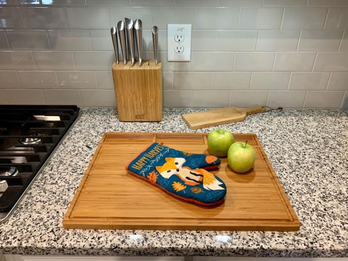 A kitchen counter showcasing a cutting board, complete with an oven mitt featuring a fox design and two green apples, with a knife block in the background.