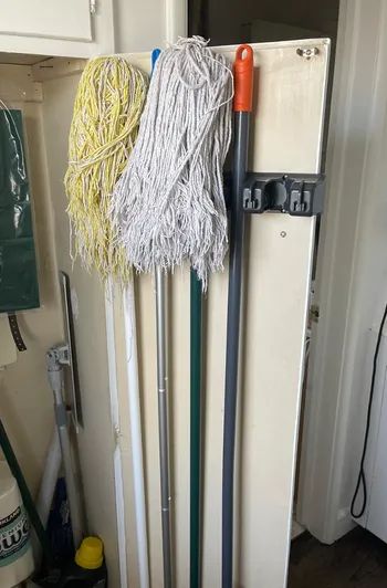 Three mops, one yellow and two white, hanging on a utility rack beside a red-handled broom in a storage area, with cleaning supplies visible on the left.