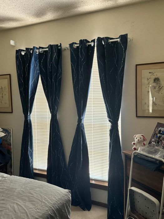 Three tall windows adorned with dark, patterned curtains tied back, allowing sunlight to stream into a cozy bedroom featuring framed artwork and a plush toy resting on a chair.