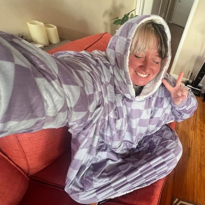 A person relaxing on a sofa in an oversized hooded blanket, flashing a peace sign, in a cozy home environment.