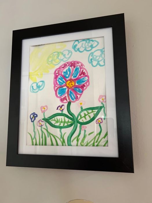 A framed child’s drawing of a large flower with a smiling center, surrounded by smaller flowers and clouds.