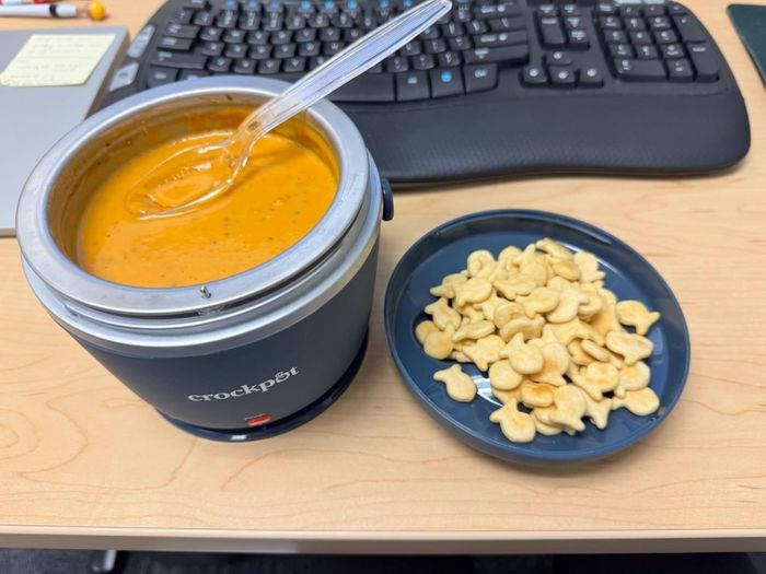 A Crockpot filled with soup, accompanied by a bowl of goldfish crackers on an office desk, with a keyboard in the background