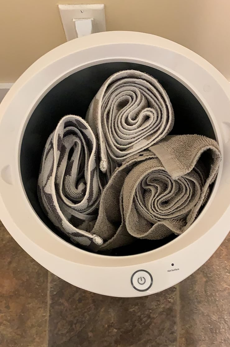 A laundry basket filled with neatly rolled towels, viewed from above, with a pair of feet visible on the floor next to the basket