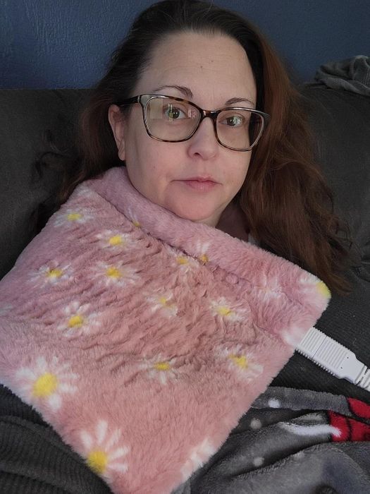 A person wearing glasses rests on a couch with a floral-patterned heating pad draped over their shoulder.