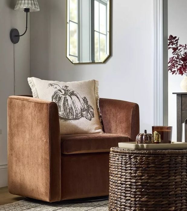 A cozy living room featuring a brown velvet chair, a pumpkin-themed pillow, a wicker table, candles, and autumn decor, all mirroring the current seasonal trends in home decor.