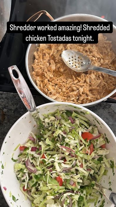 A pan filled with perfectly shredded chicken alongside a slotted spoon. Nearby, a bowl brimming with shredded lettuce, red onion, and tomatoes. Caption: "This shredder is incredible! Made the best shredded chicken tostadas tonight."