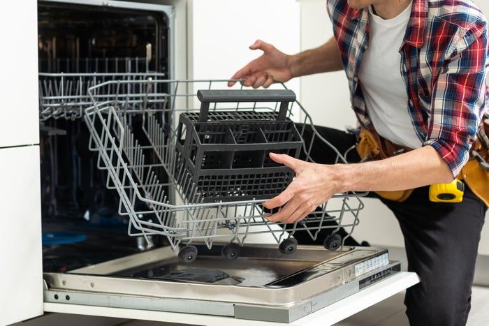 A person repairing an open dishwasher, hands on the racks, with a tool belt visible