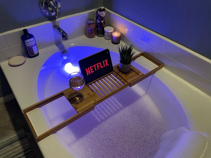 A serene bathtub setup featuring a wooden caddy, with a tablet showing Netflix, a glass of wine, lit candles, and a potted plant, creating a tranquil spa atmosphere.