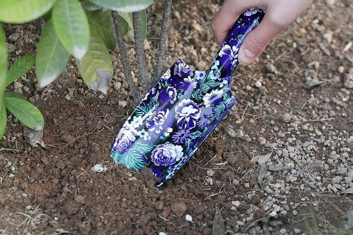 A hand gripping a floral-patterned garden trowel, carefully digging soil around a plant, showcasing gardening tools in a shopping guide