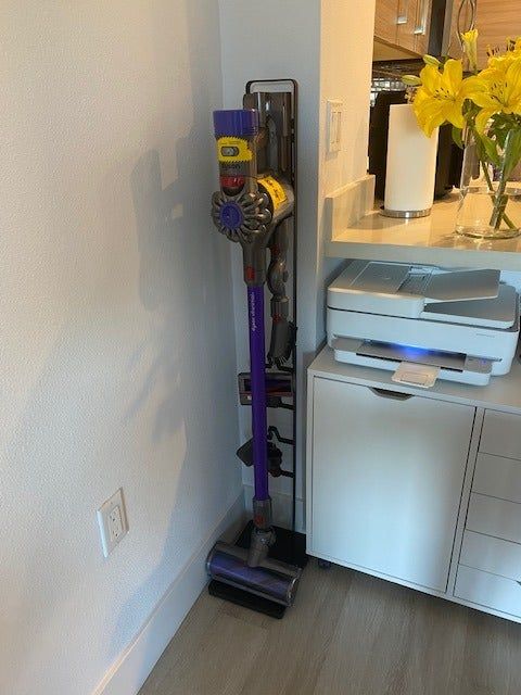 A cordless vacuum cleaner standing upright next to a printer on the floor in a home setting.