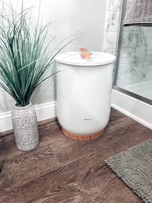 A stylish white laundry hamper with wooden accents, placed beside a decorative vase and shower in a chic bathroom setting