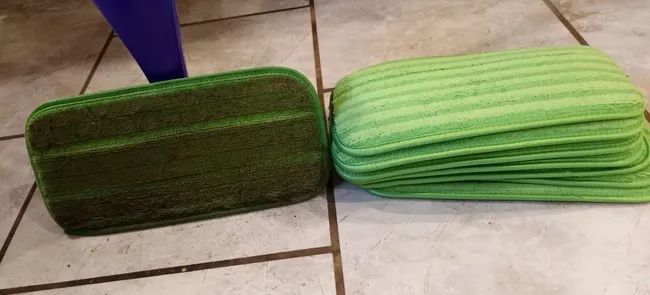 A dirty green mop head placed on a Swiffer next to clean pads stacked in a pile.
