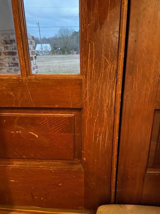 A wooden door showing visible scratch marks, with a rural landscape visible through the window and part of a chair in the lower corner.