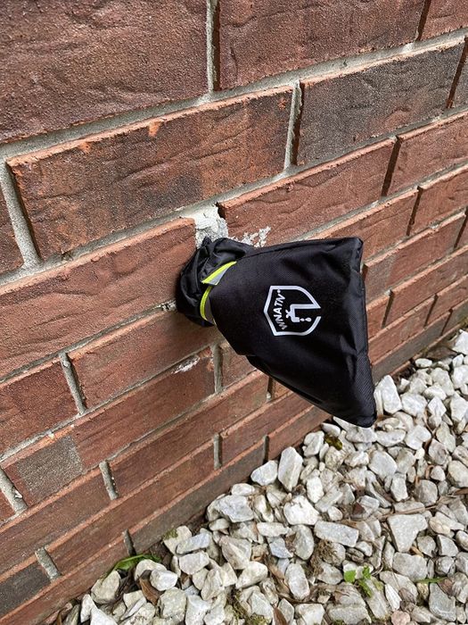 A black insulated cover on an outdoor water spigot attached to a brick wall, with a white logo visible on the cover. Gravel is seen below the wall