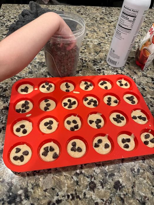 A hand gently arranges chocolate chips onto balls of cookie dough in a red baking tray, ready for baking.