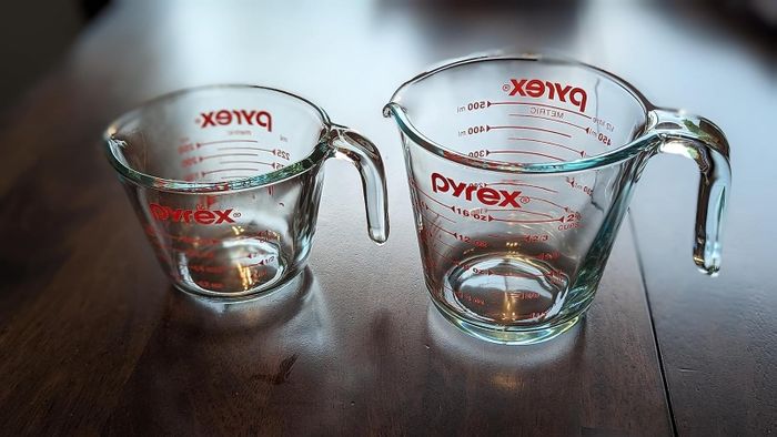 Two Pyrex glass measuring cups—one large, one small—placed on a rustic wooden table, both marked with red measurement indicators.
