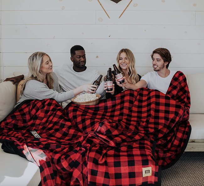 Four models cozily bundled up inside a large red and black checkered blanket on the couch