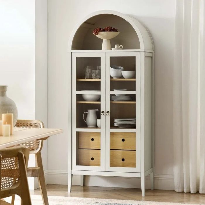 A sophisticated cabinet featuring an arched top, glass-paneled doors, and wooden drawers. Styled with elegant tableware inside, making it a perfect statement piece for home decor enthusiasts.