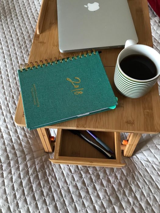 A notebook, laptop, and coffee placed on a wooden tray with a small drawer holding pens, positioned on a quilted surface