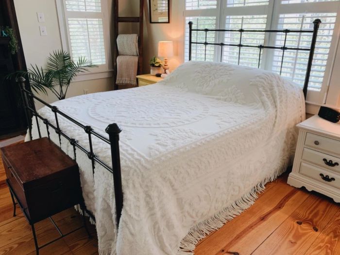 A bedroom featuring a bed with a detailed white bedspread, wooden flooring, and a plant in the corner for a warm, inviting atmosphere