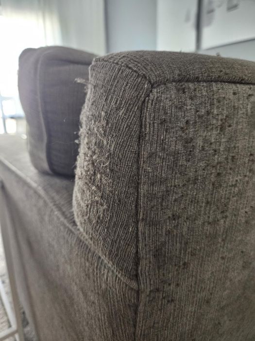 A close-up view of a worn corner on a grey couch, showing clear signs of fabric fraying and pilling.