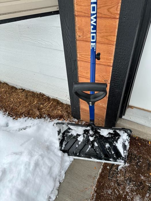 A snow shovel rests against a wall near a snow-covered walkway.