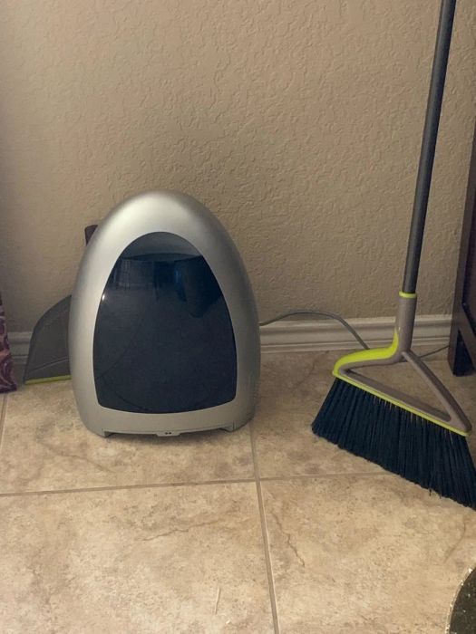 The gray and black bagless, touchless vacuum resting on a tiled floor beside a broom