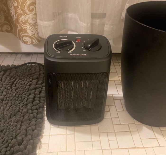 A portable space heater placed on a tiled floor next to a textured rug and a trash can. The heater comes with adjustable settings and a caution label for safety.